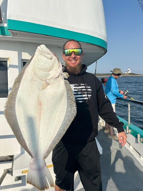 Man holding a large fish on a boat with a scenic ocean view and lighthouse in background.