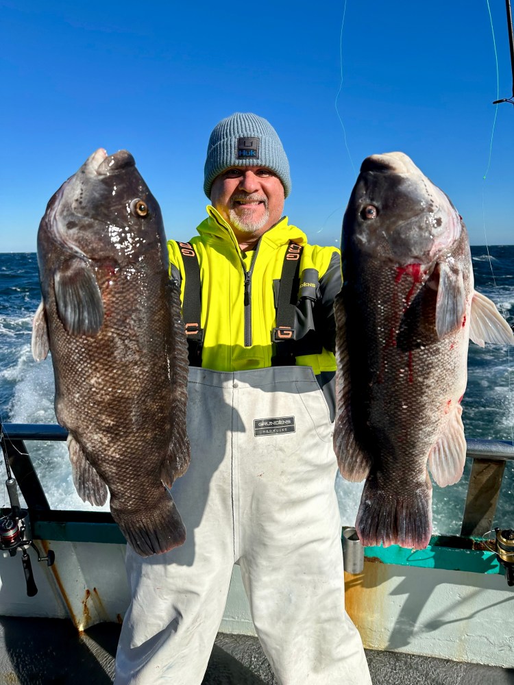 Person in yellow jacket holding two large fish on a boat with ocean background.