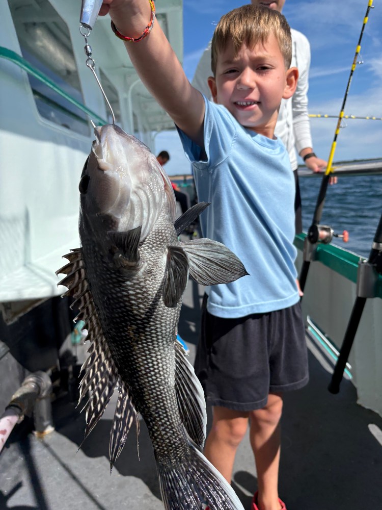 Child on a boat proudly holding a large fish by a line under a clear blue sky.
