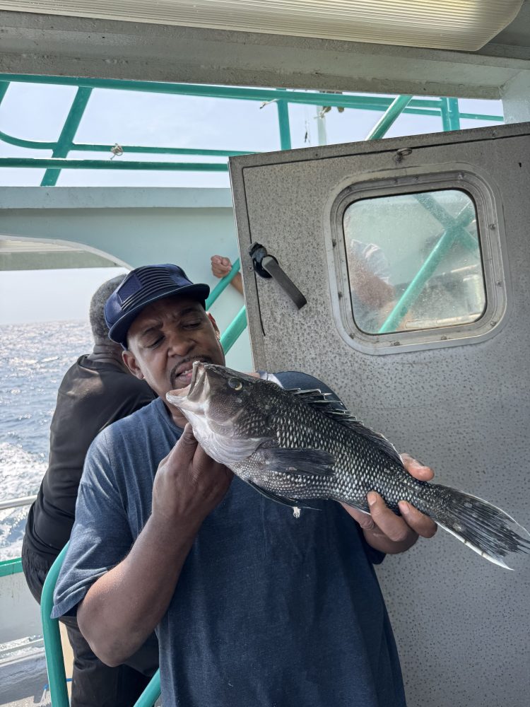 Man on a boat playfully poses with a large fish near his face.