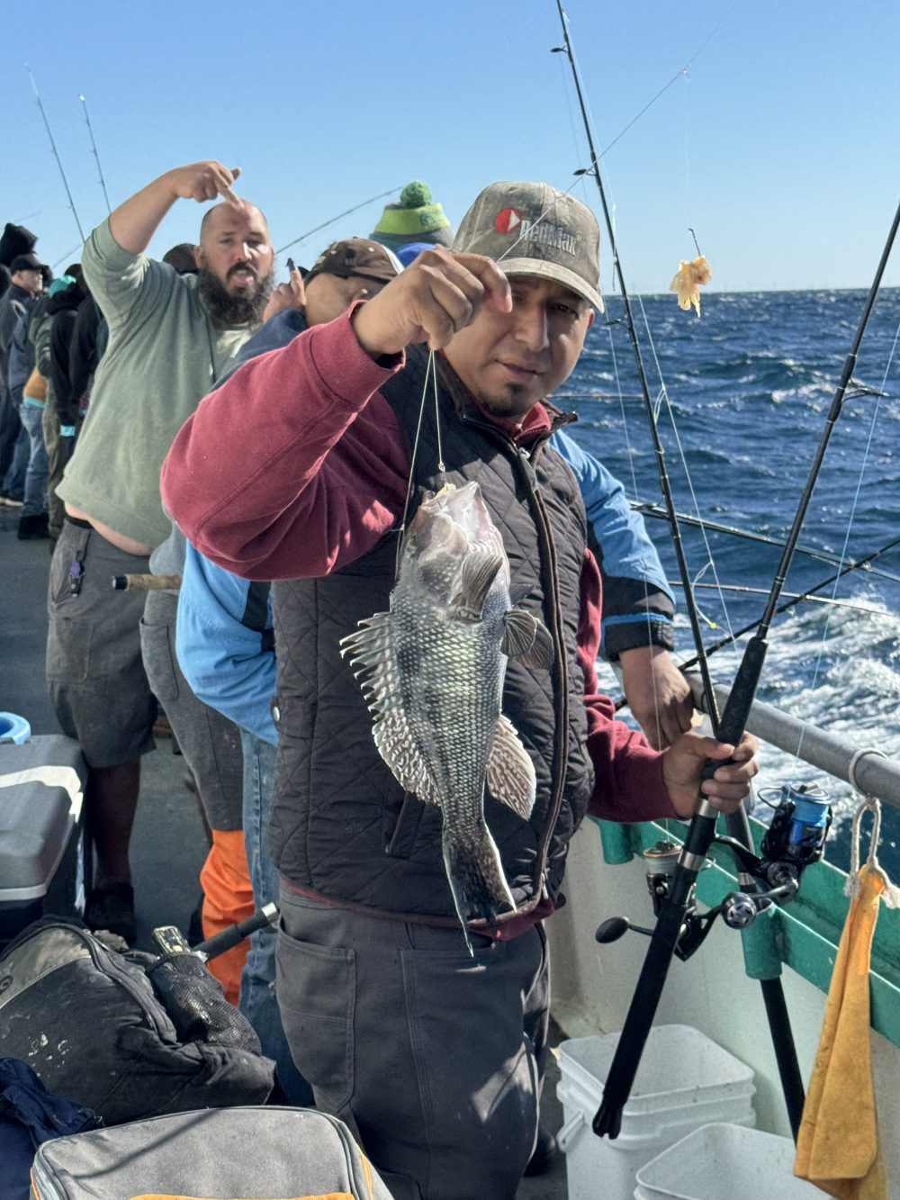 Man on boat holding a fish with fishing pole, ocean in background and people fishing.