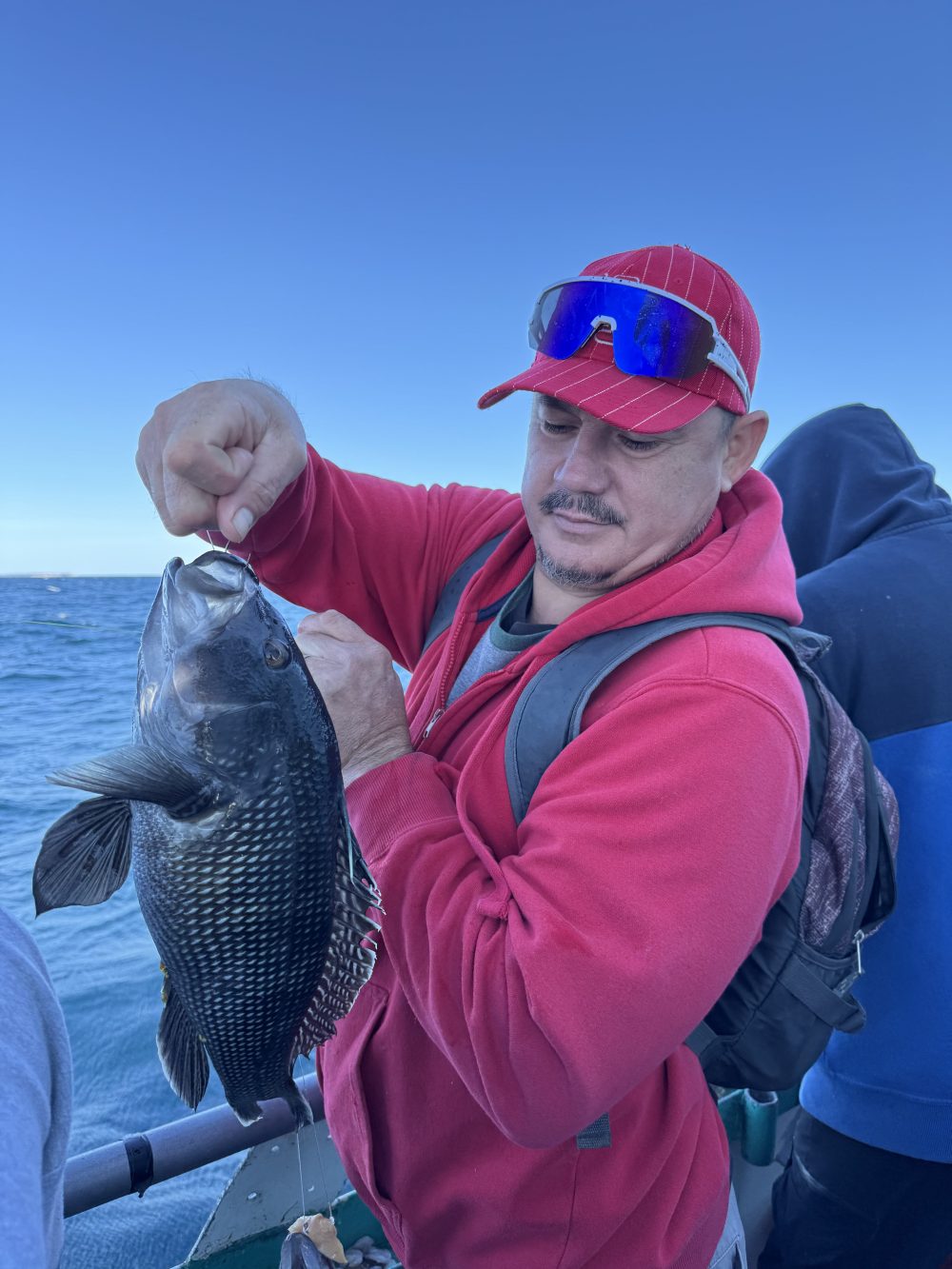 Person in red hoodie and cap holding a fish on a boat with ocean background.