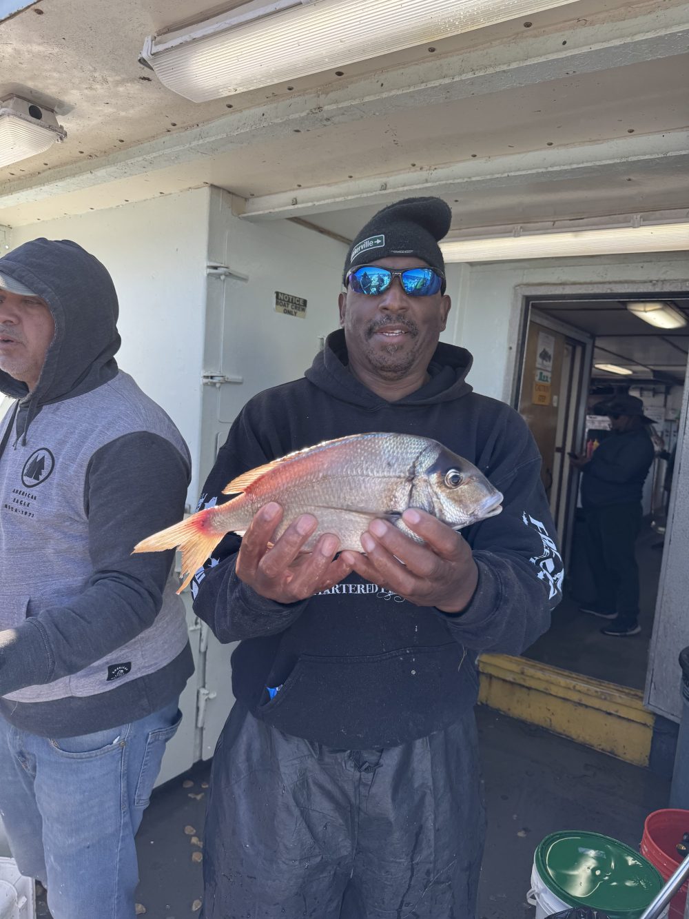 Man smiling, wearing sunglasses, holding a fish on a boat with another person nearby.