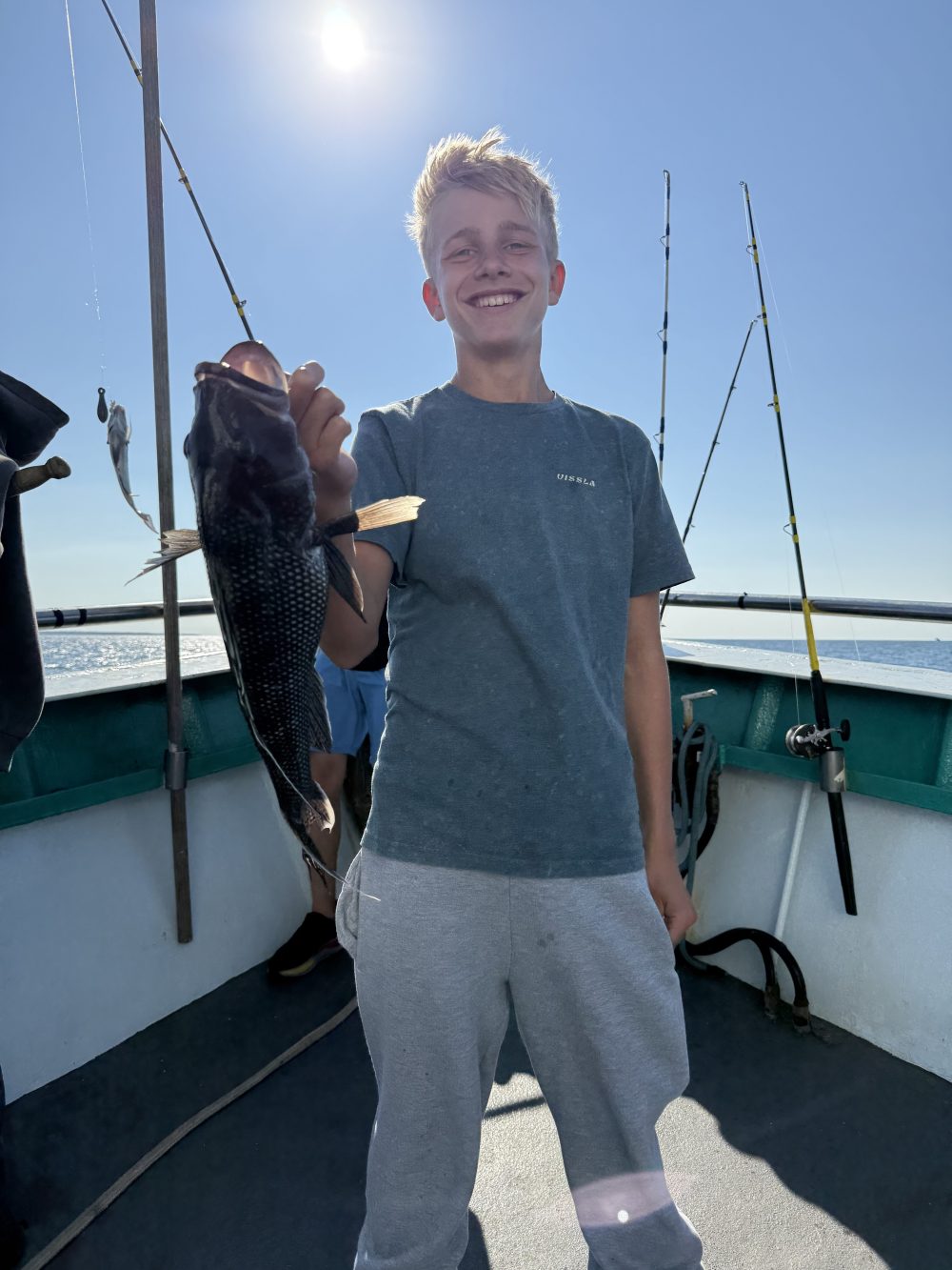 Person smiling while holding a fish on a boat with fishing rods in the background under a sunny sky.