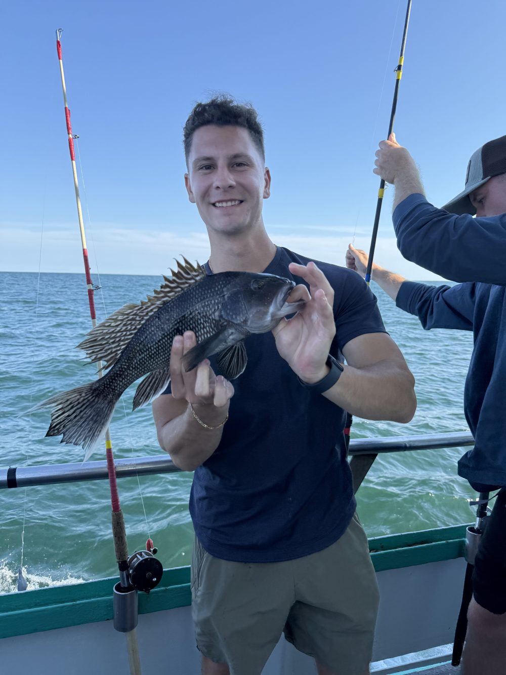 Person holding a fish on a boat with fishing rods and ocean in the background.