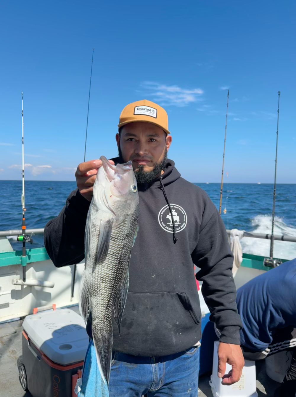 Man holding a large fish on a boat with blue sky and sea in the background.