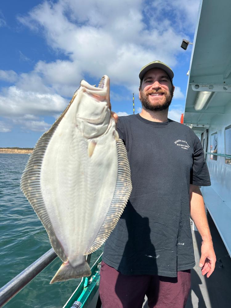 Man on a boat holding a large flatfish with the sea and clouds in the background.