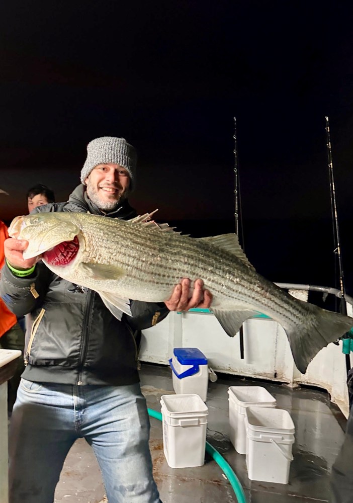 Man holding large striped bass on a boat at night, wearing a beanie and jacket.