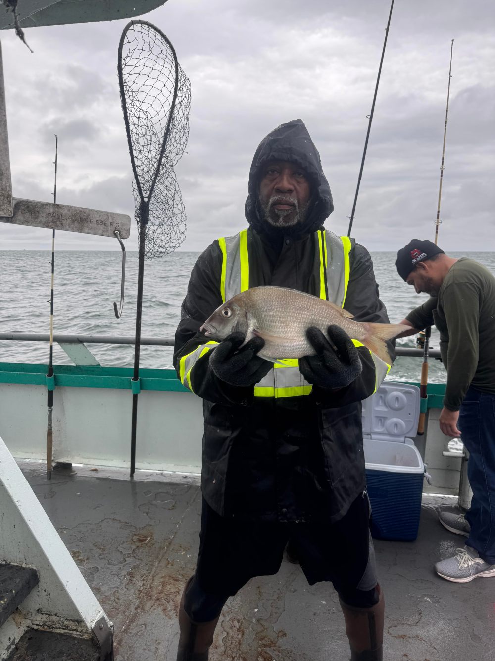 Person in rain gear holds a fish on a boat with overcast sky.