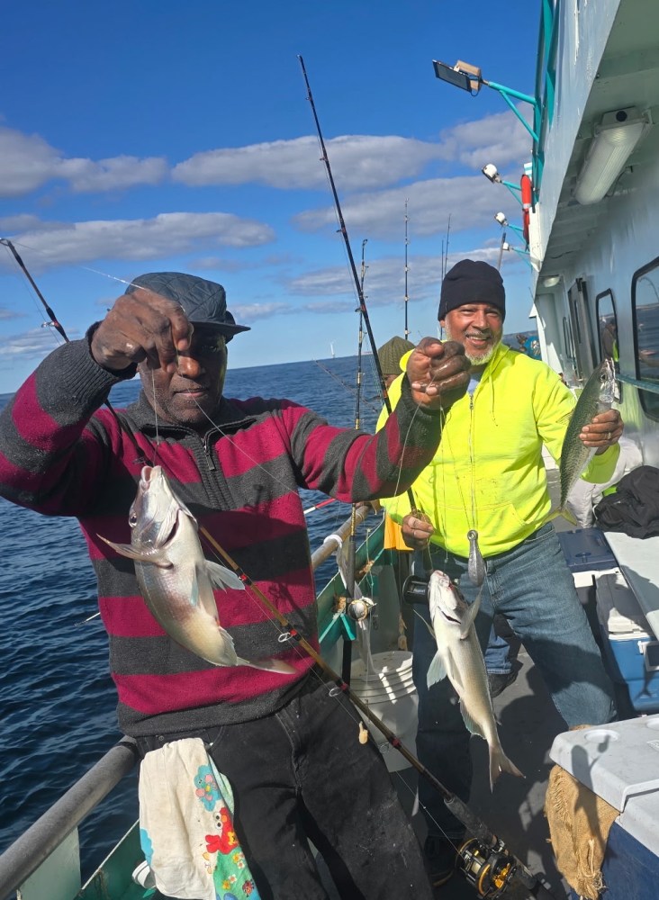 Two men on a boat holding fishing lines with several caught fish, smiling with blue sky and sea background.