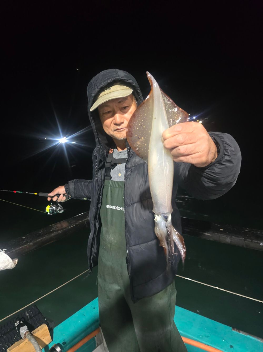 Person holding a large squid on a fishing boat at night with bright lights in the background.