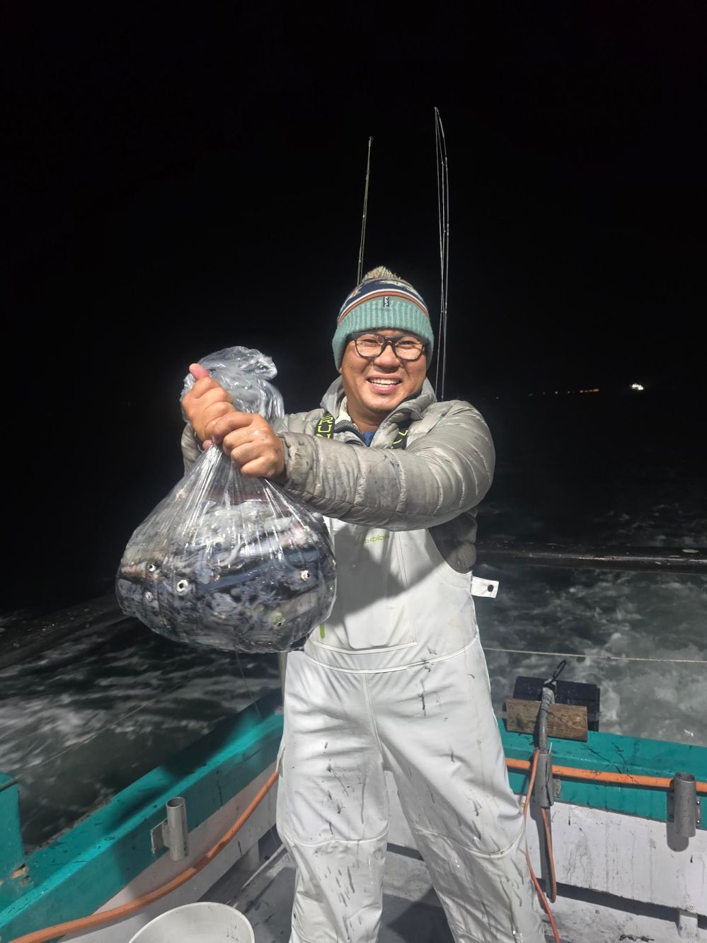 Person on a boat at night holding a bag of fish, wearing a beanie and overalls, smiling.
