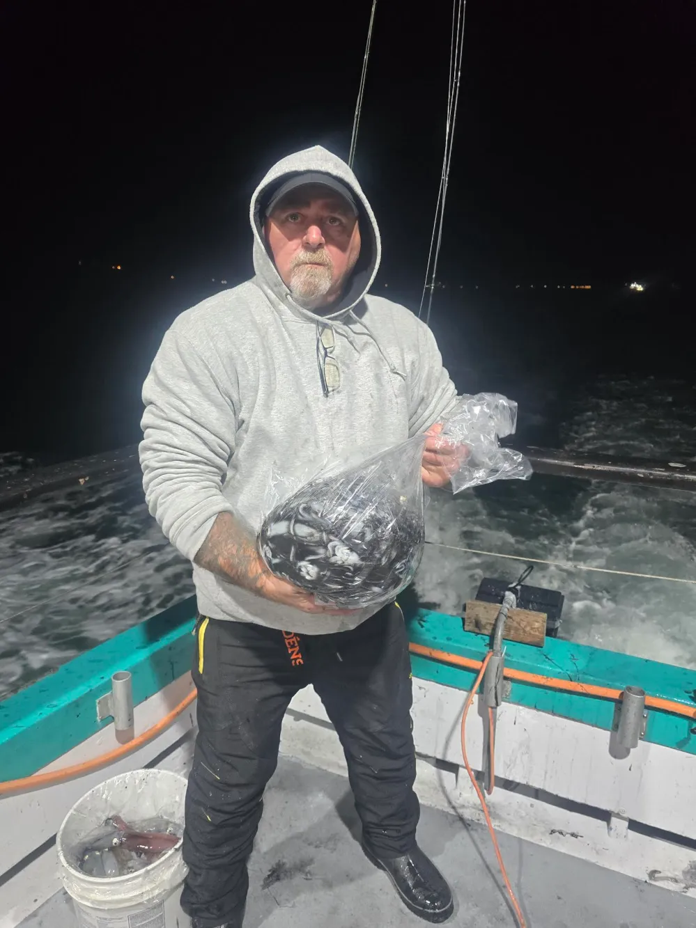 Man in hoodie holding bag of fish on a boat at night.