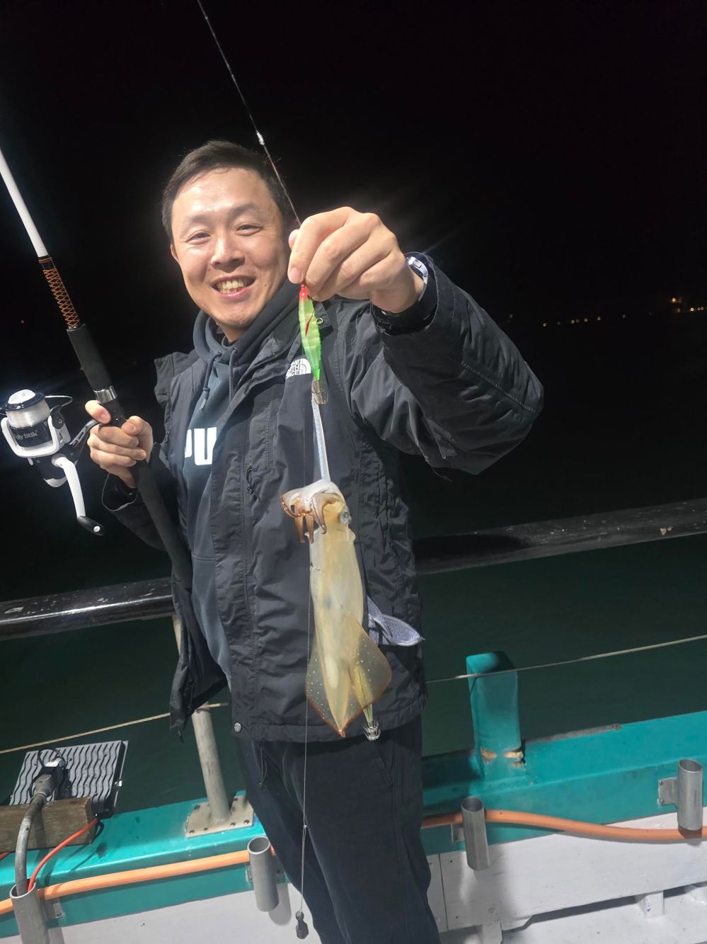 Person smiling, holding a fishing rod with a squid on a boat at night.