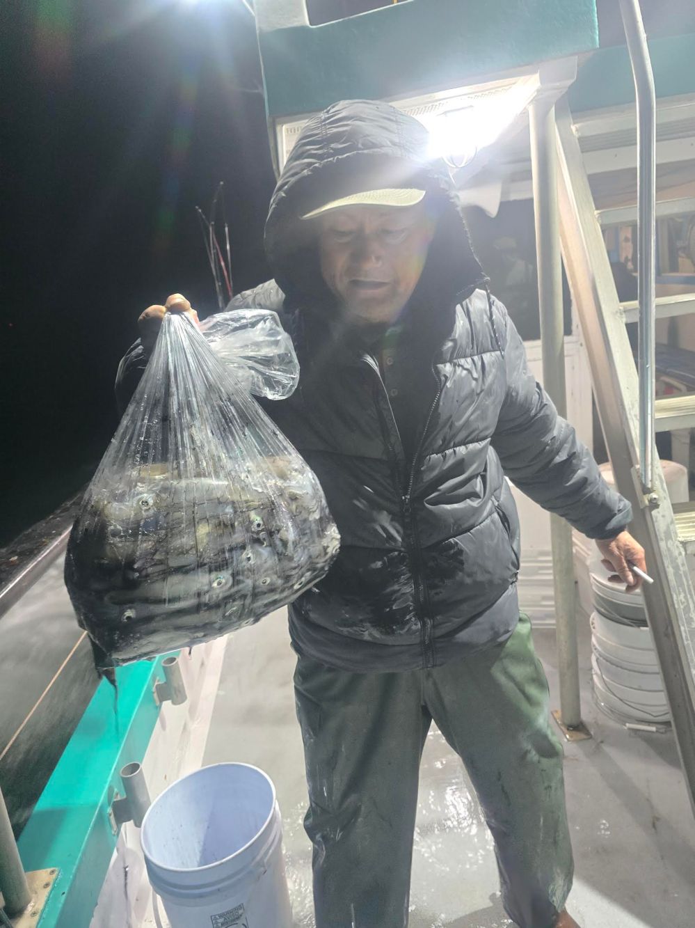 Person in a hooded jacket on a boat holding a bag of fish at night.