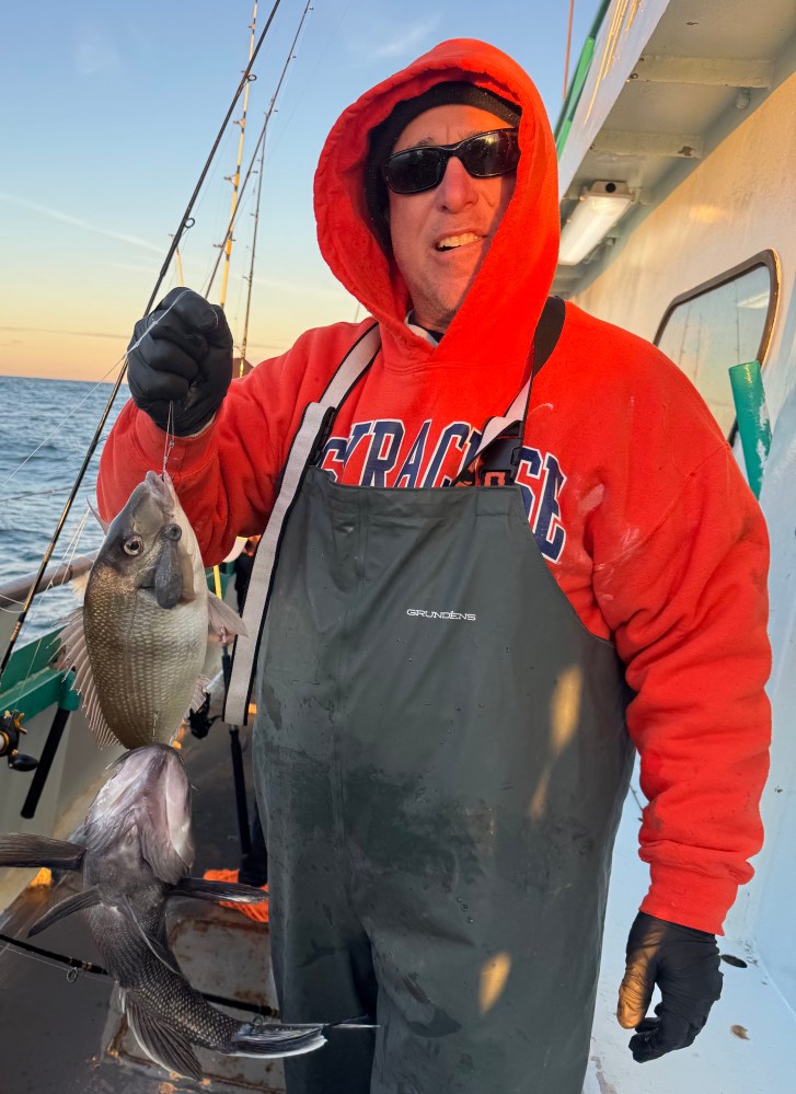 Person in red hoodie holding two fish on a boat at sunset.