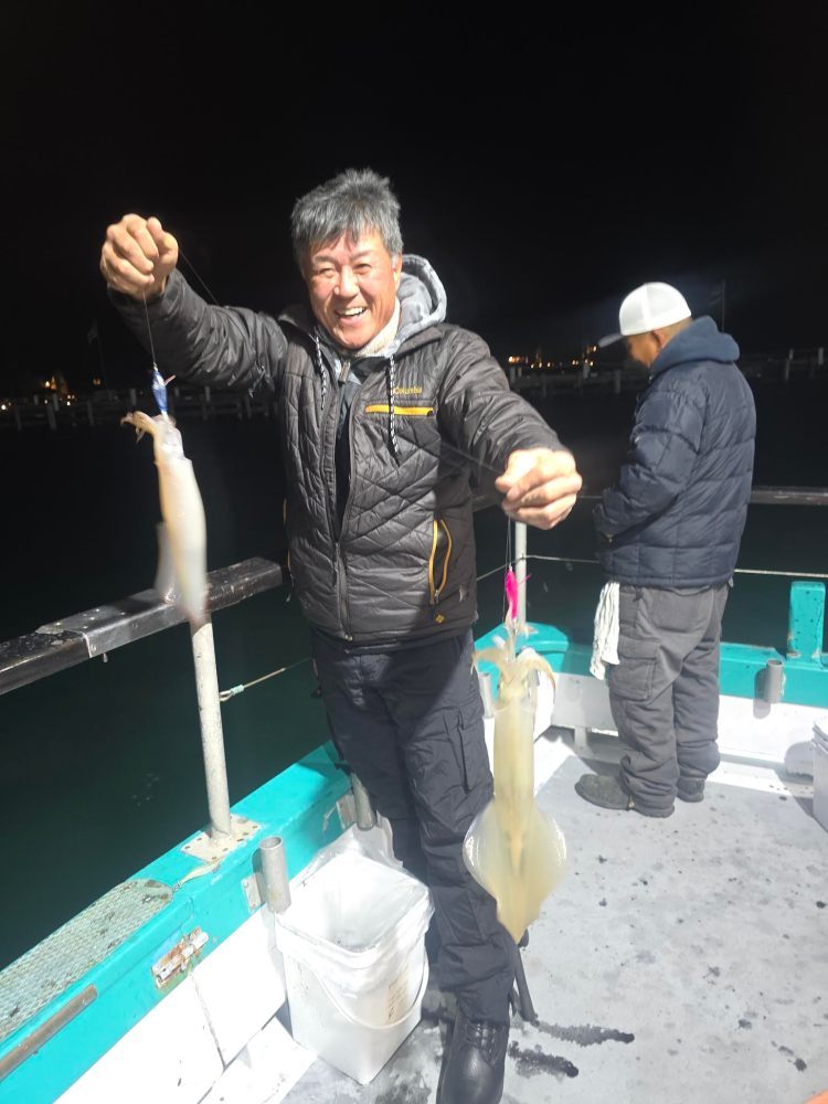 Man smiling, holding two squids on a boat at night, another person facing away.
