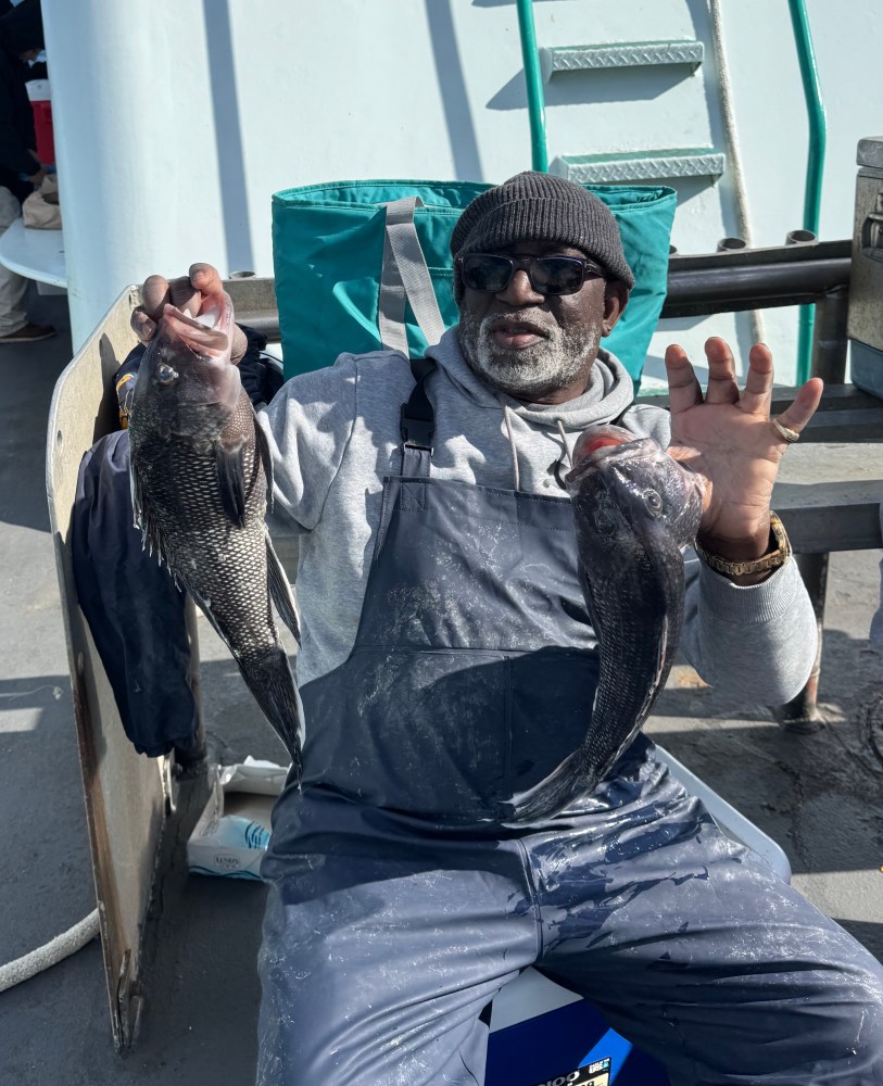 Man in sunglasses holding two fish on a boat, smiling, wearing a beanie and overalls.