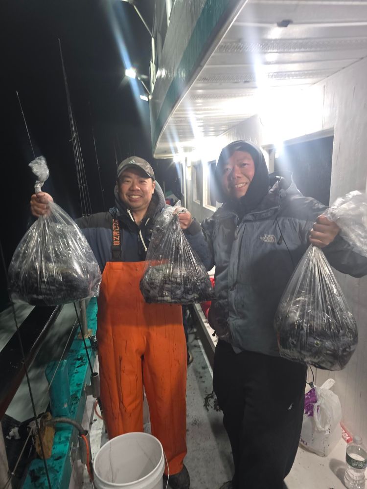 Two men on a boat holding bags of freshly caught fish at night.