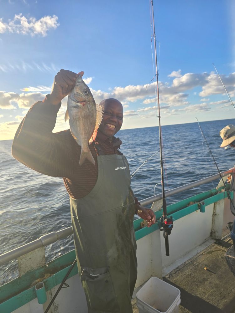 Man on boat holding a fish with a smiling face, ocean and fishing rods in background.