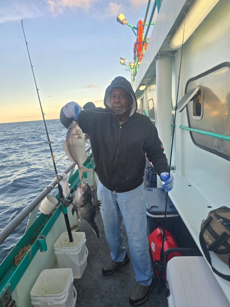 Person in a hoodie holding two fish on a boat at sea with fishing gear visible.