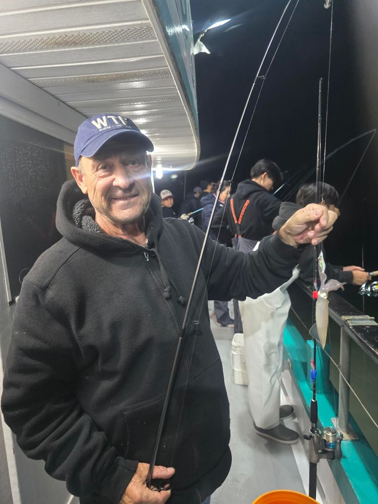 Man in a black hoodie and cap holding a fishing rod on a boat at night.