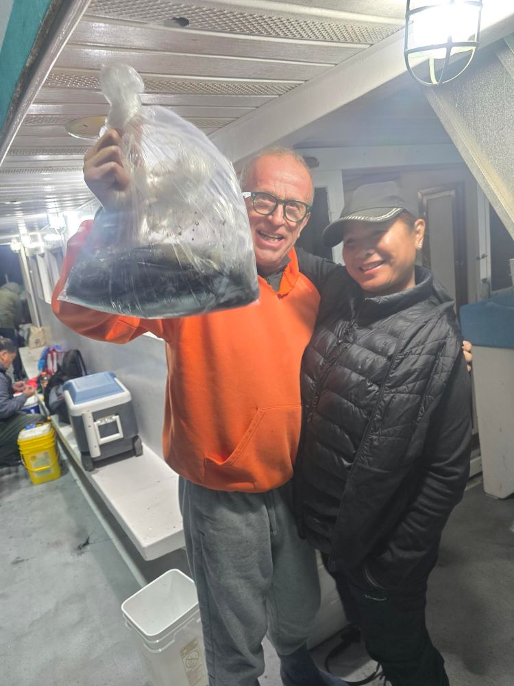 Two people on a boat, one holding a bag of fish, smiling at the camera.