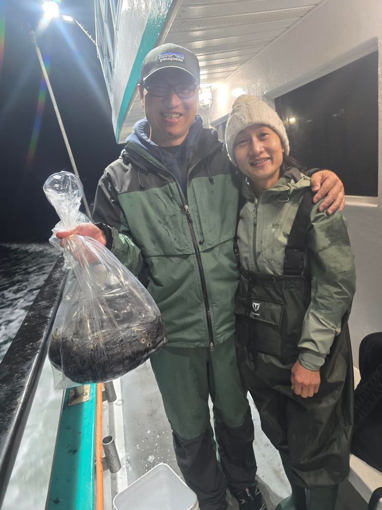 Two people in rain gear on a boat at night, one holding a plastic bag with a fish.