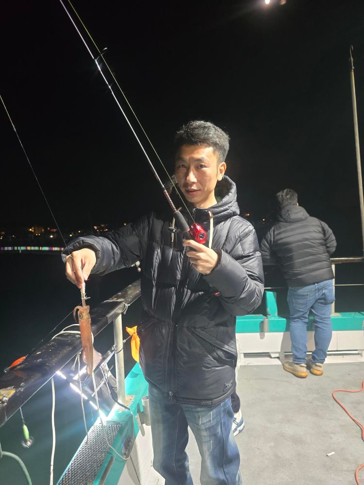 Man in a puffy jacket fishing at night on a boat, holding a squid on a line.