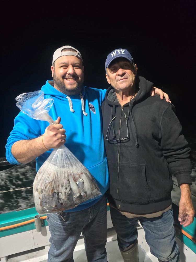 Two men smiling on a boat with one holding a bag of fish.