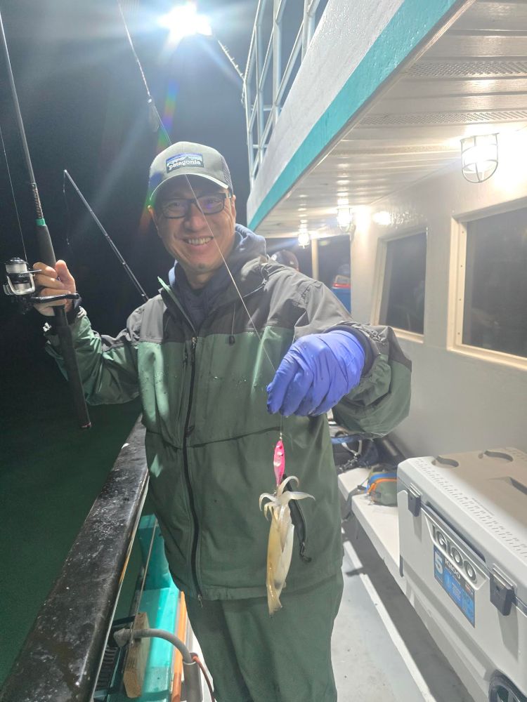 Person in outdoor gear holding a fishing rod with a squid on a boat at night.