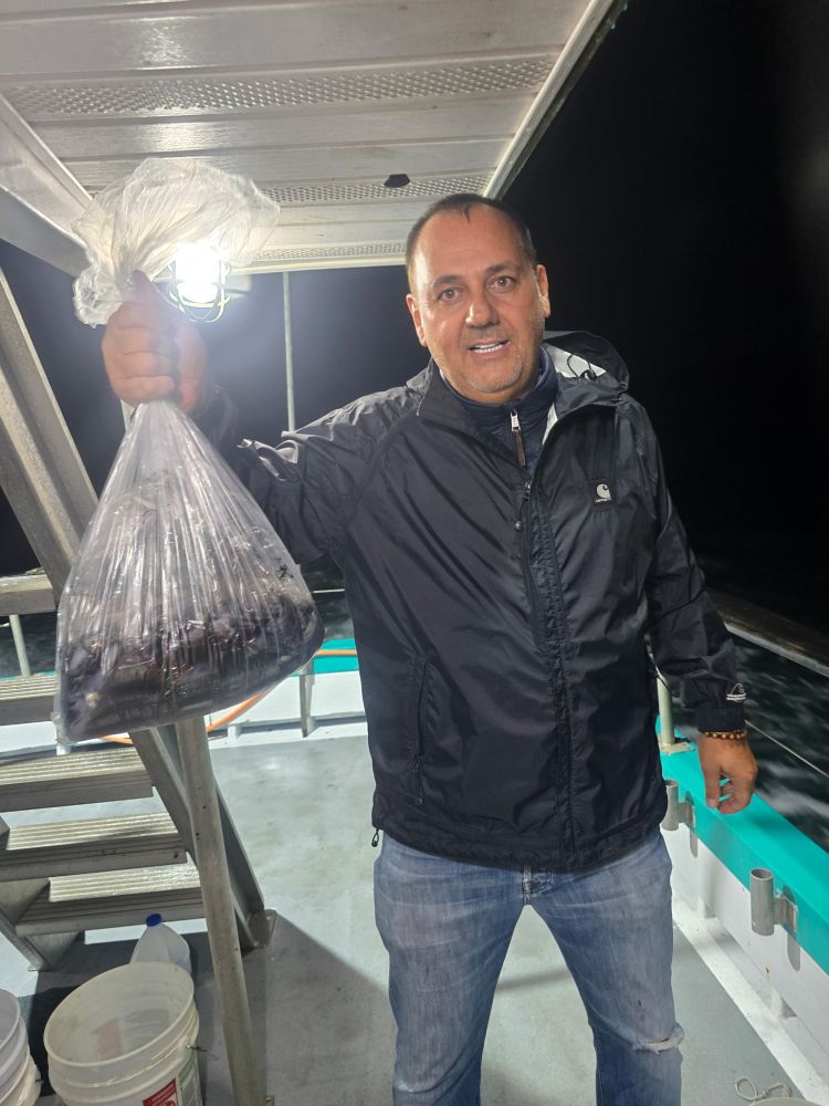 Man on a boat holding a plastic bag filled with fish during nighttime.
