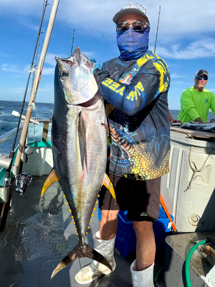 Person on a boat holding a large fish, wearing a face cover and sunglasses on a sunny day.