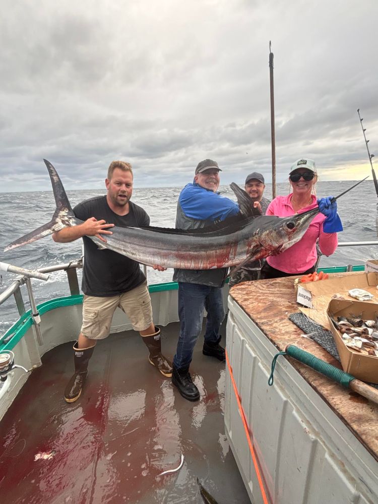 Four people on a boat holding a large fish with the ocean in the background.