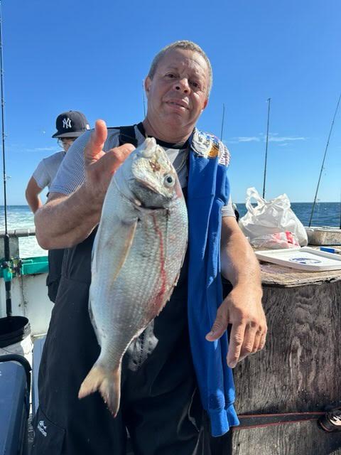 Man on boat holding a large fish with ocean and fishing rods in background.