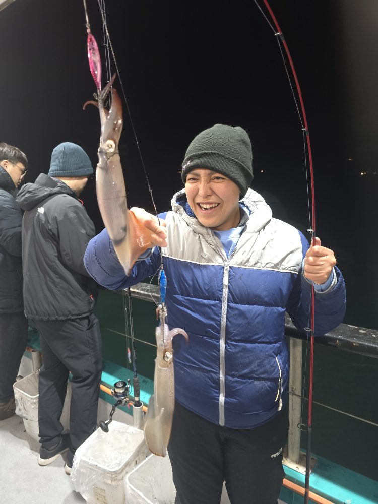Person on a boat holding a fishing rod with two caught squids, smiling and wearing a beanie.