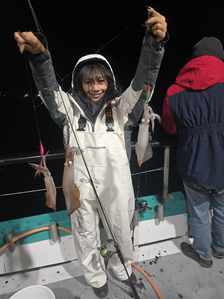 Person in white fishing suit holds two squids on a boat at night.