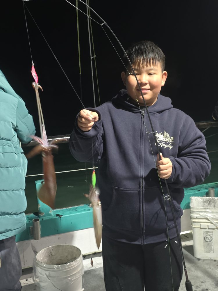 Boy on boat holds fishing rod with squid at night, smiling.