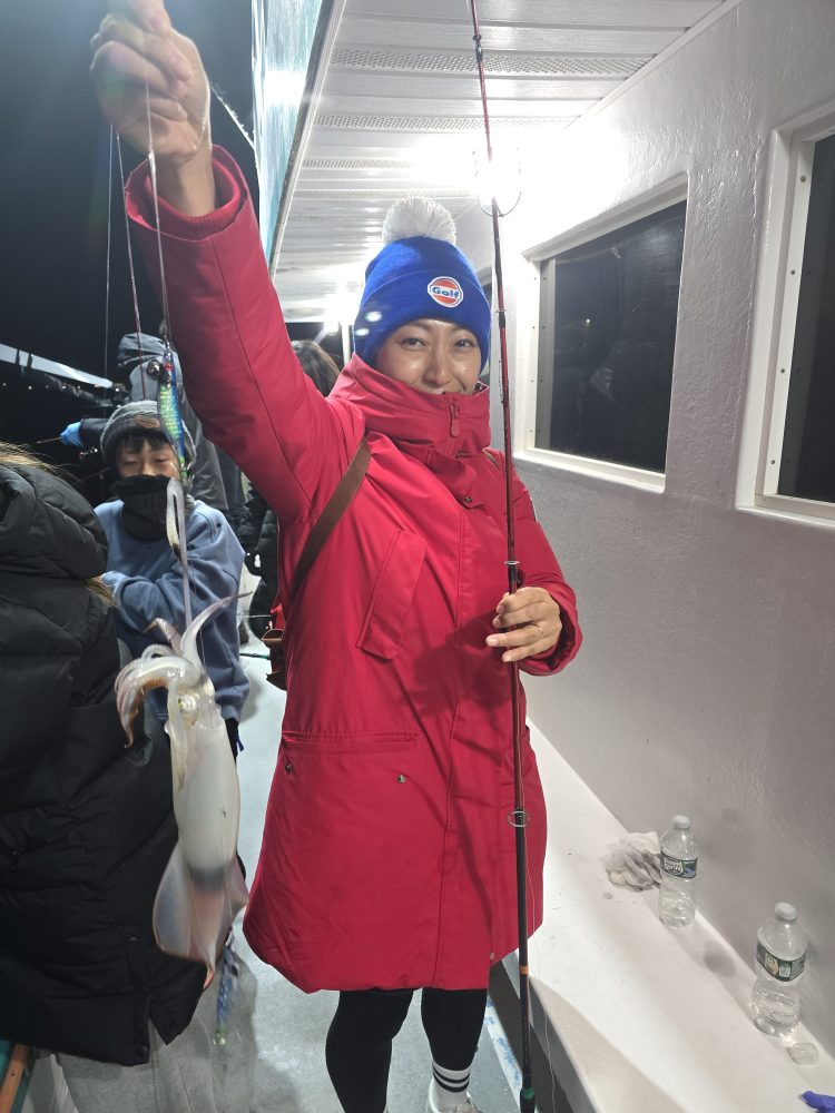 Person in red coat and blue hat holding a squid on a fishing line on a boat at night.