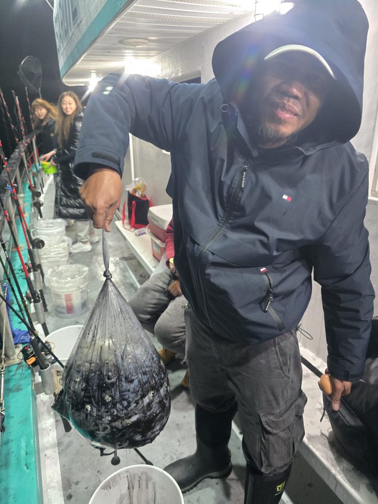 Person holds net bag with fish on a boat at night, with fishing rods in the background.
