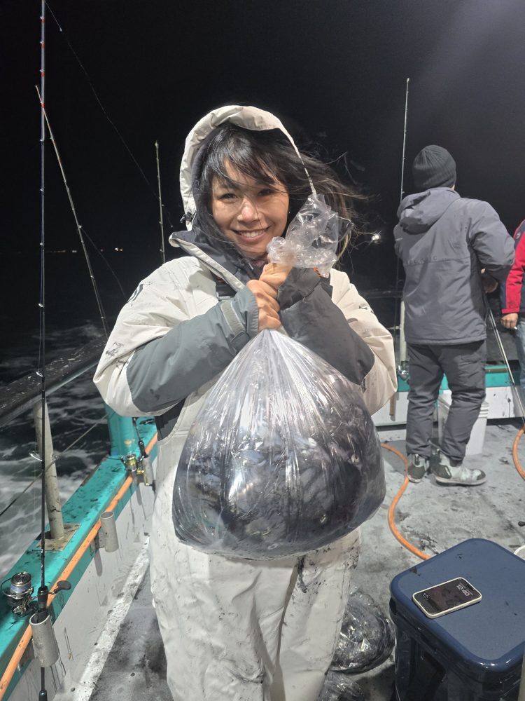 Person in a hooded jacket holds a sealed plastic bag of fish on a boat at night.