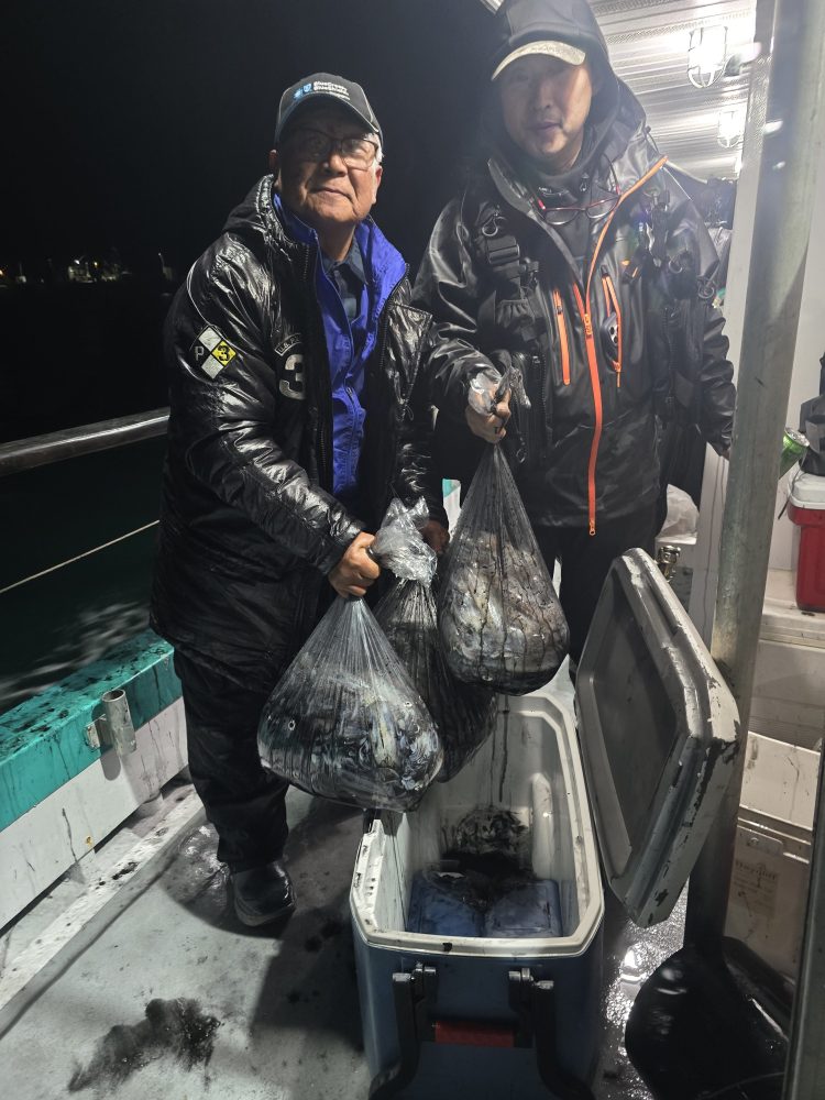 Two people in jackets holding bags of fish on a boat at night near a cooler.