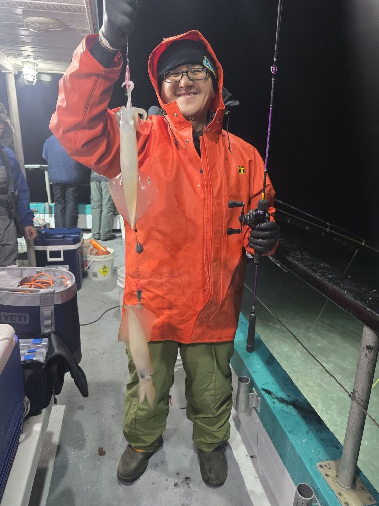 Person in orange jacket holding a squid on a fishing boat at night.