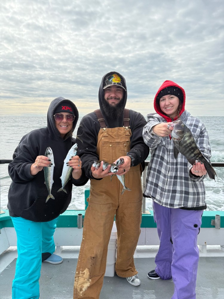 Three people on a boat holding fish, dressed warmly under a cloudy sky.