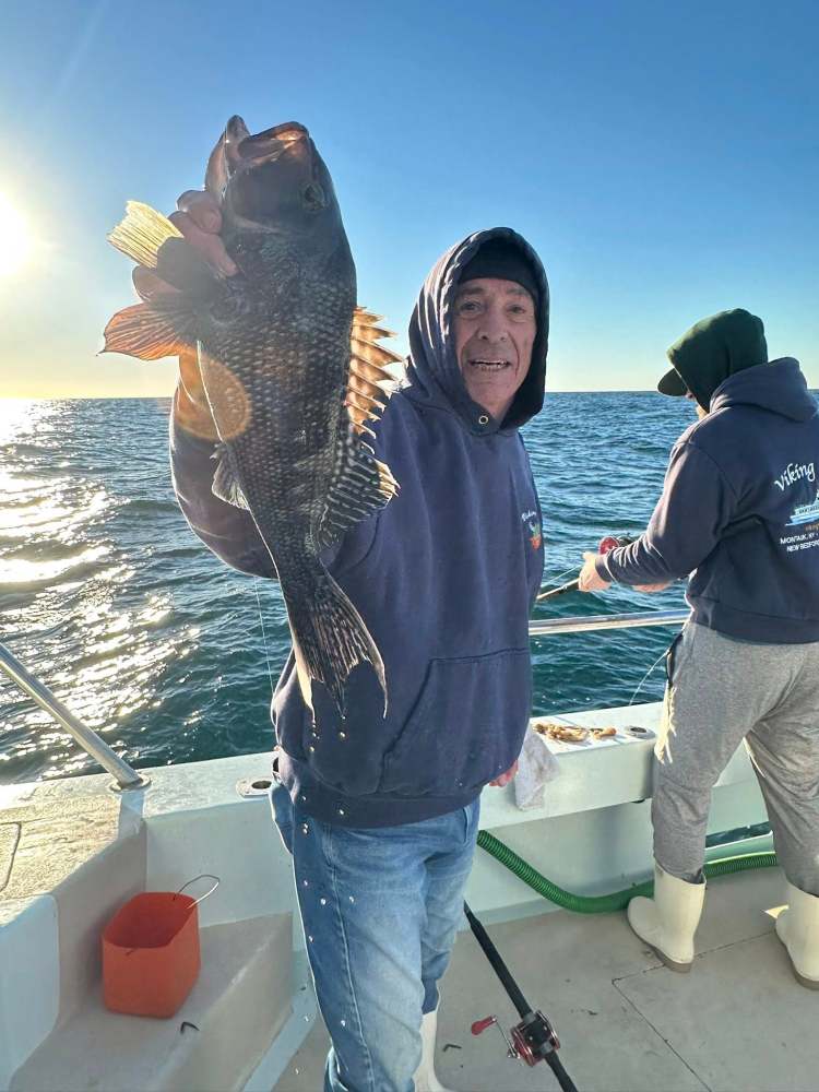 Person holding a large fish on a boat with another person fishing in the background during sunset.