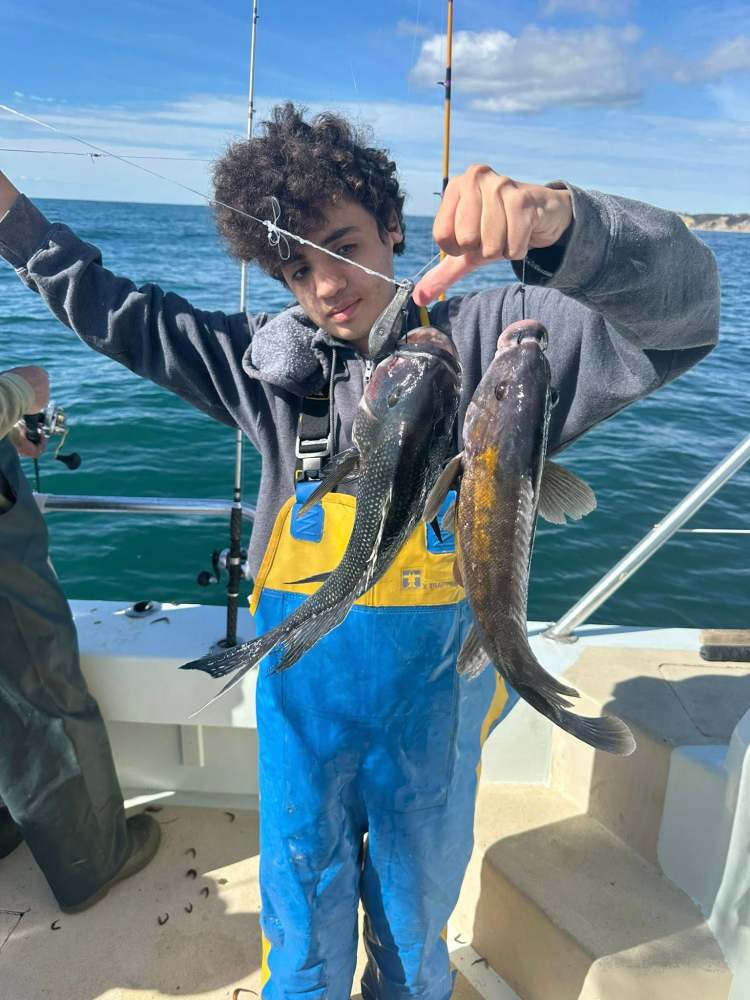Person in yellow waders holding two fish on a boat at sea.