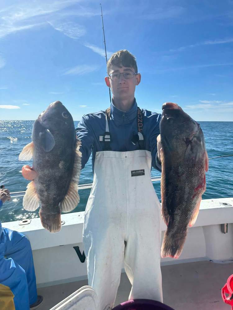 Person in white overalls holds two fish on a boat with ocean in background.