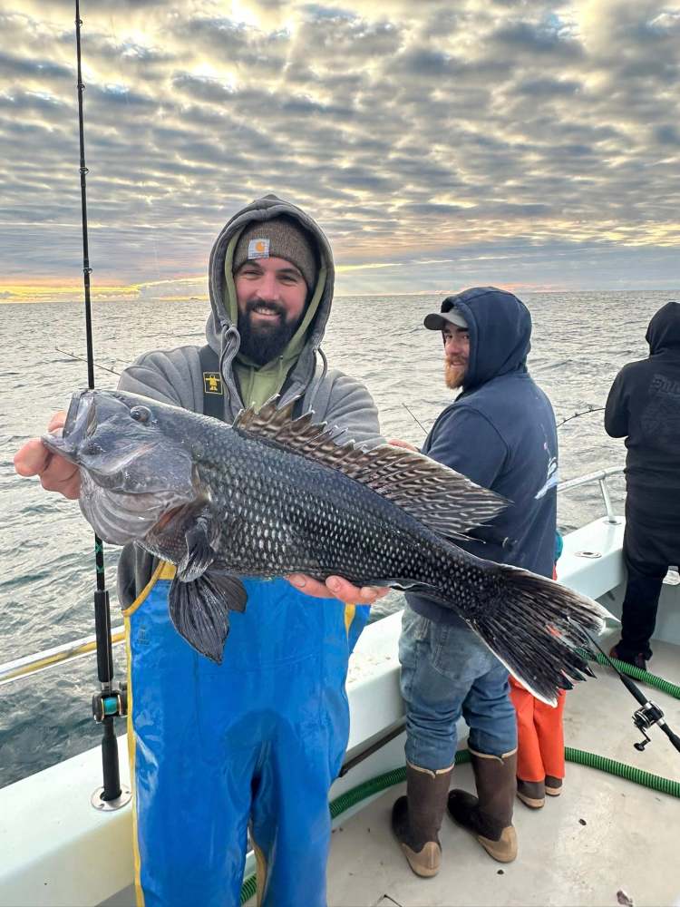 Man on boat holding a large fish, wearing blue overalls and a hooded sweatshirt.