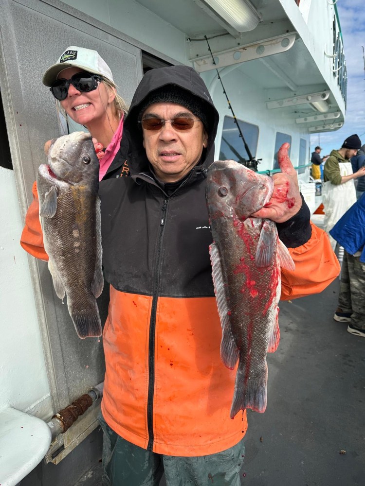 Person holding two large fish on a boat, wearing an orange jacket and black hood.
