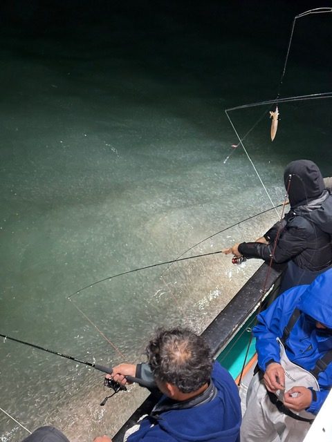 Two people fishing at night on a boat, with submerged fish visible in the water.